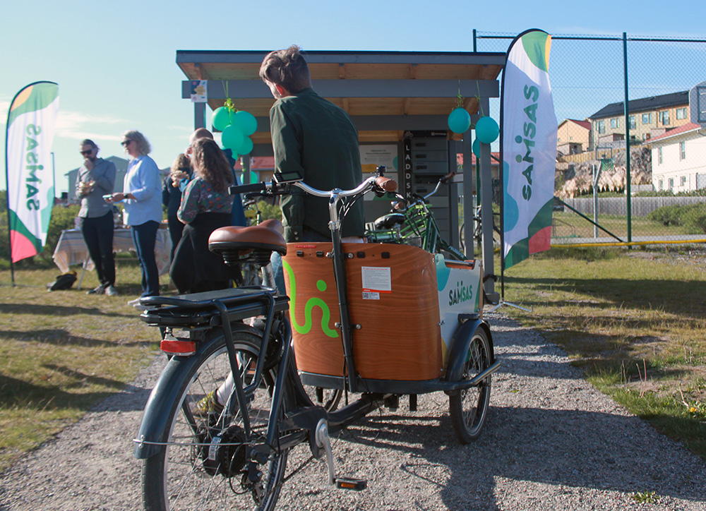 I förgrunden en lådcykel framför ett cykelgarage, intill står flera personer och lyssnar på talare.