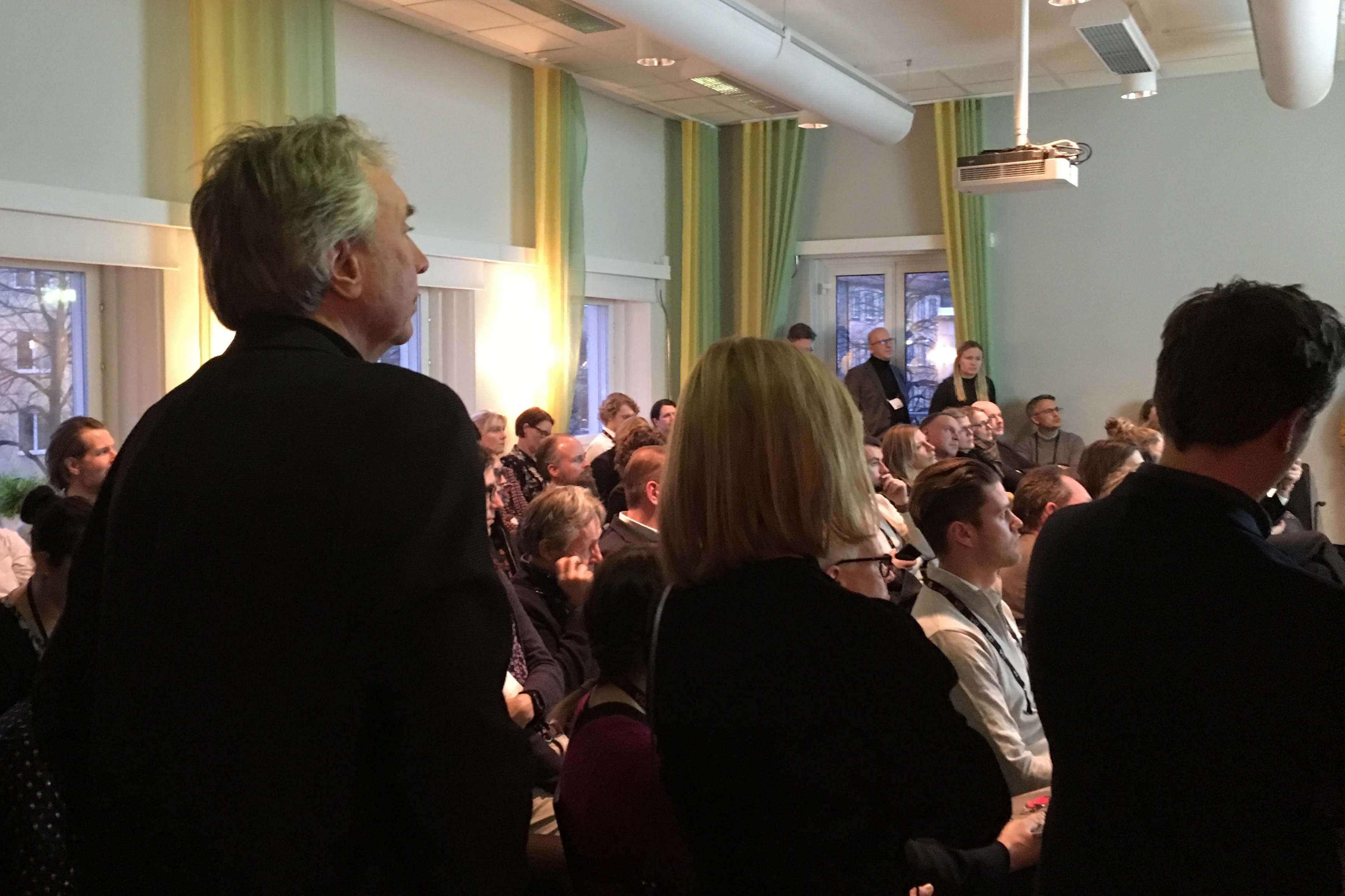 The backs of a number of people are seen through a doorway, the room within is full with seated people watching a presentation out of view from the camera.